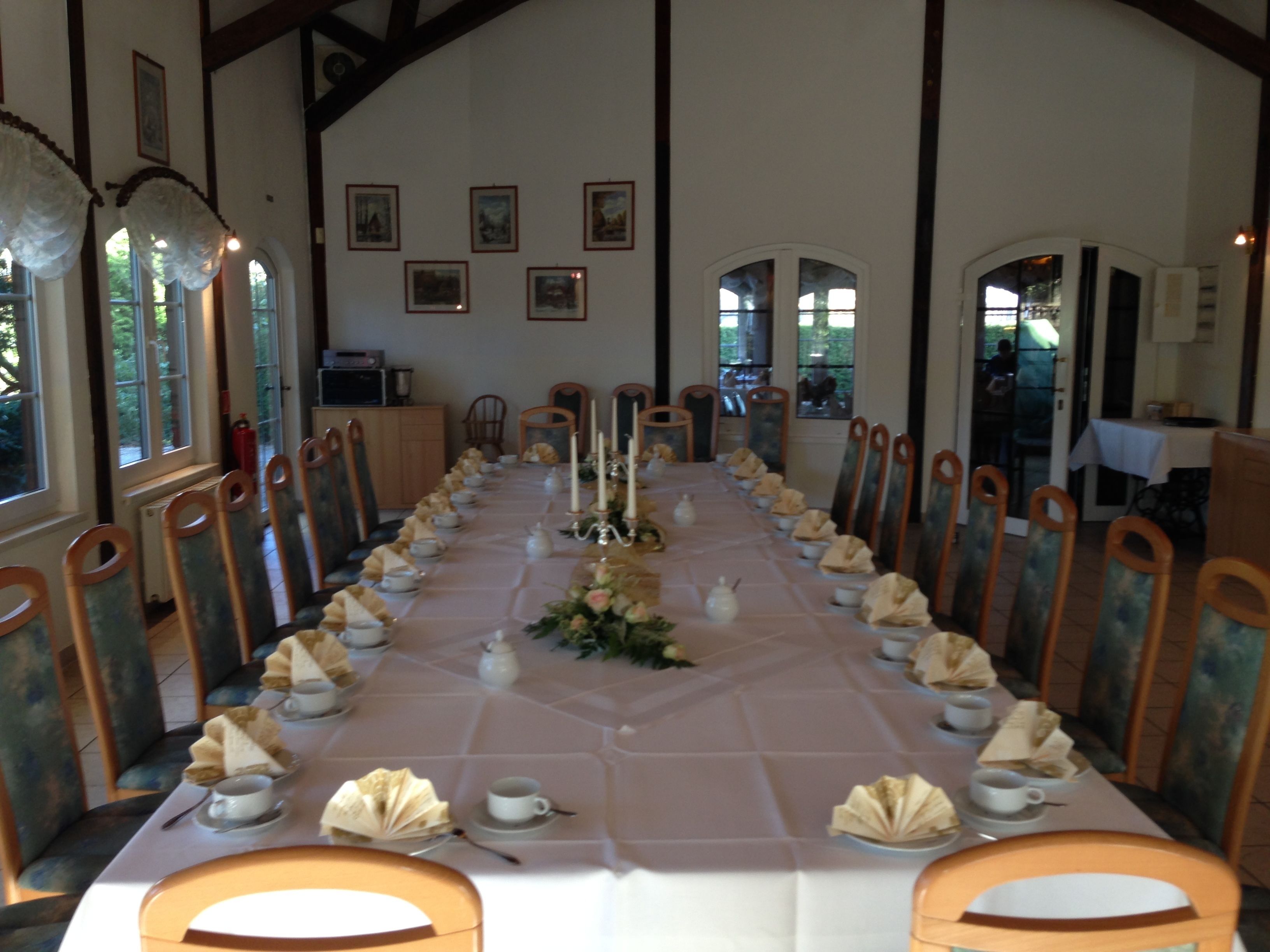 A long dining table set for a formal meal with chairs on both sides, framed pictures on the walls, and large windows.