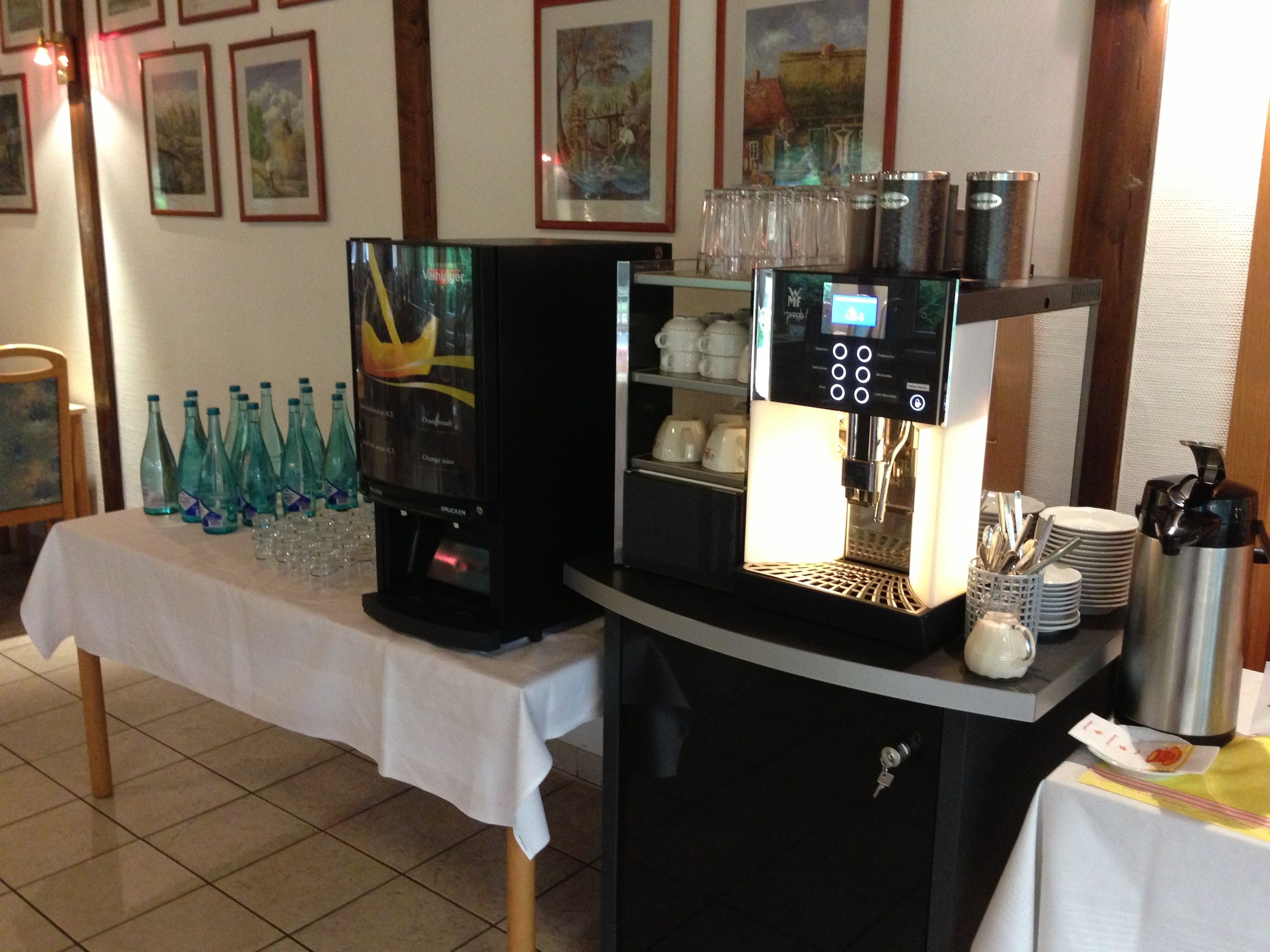 A coffee station with two espresso machines, a table with stacked cups and glasses, and a shelf with mugs and a thermos.
