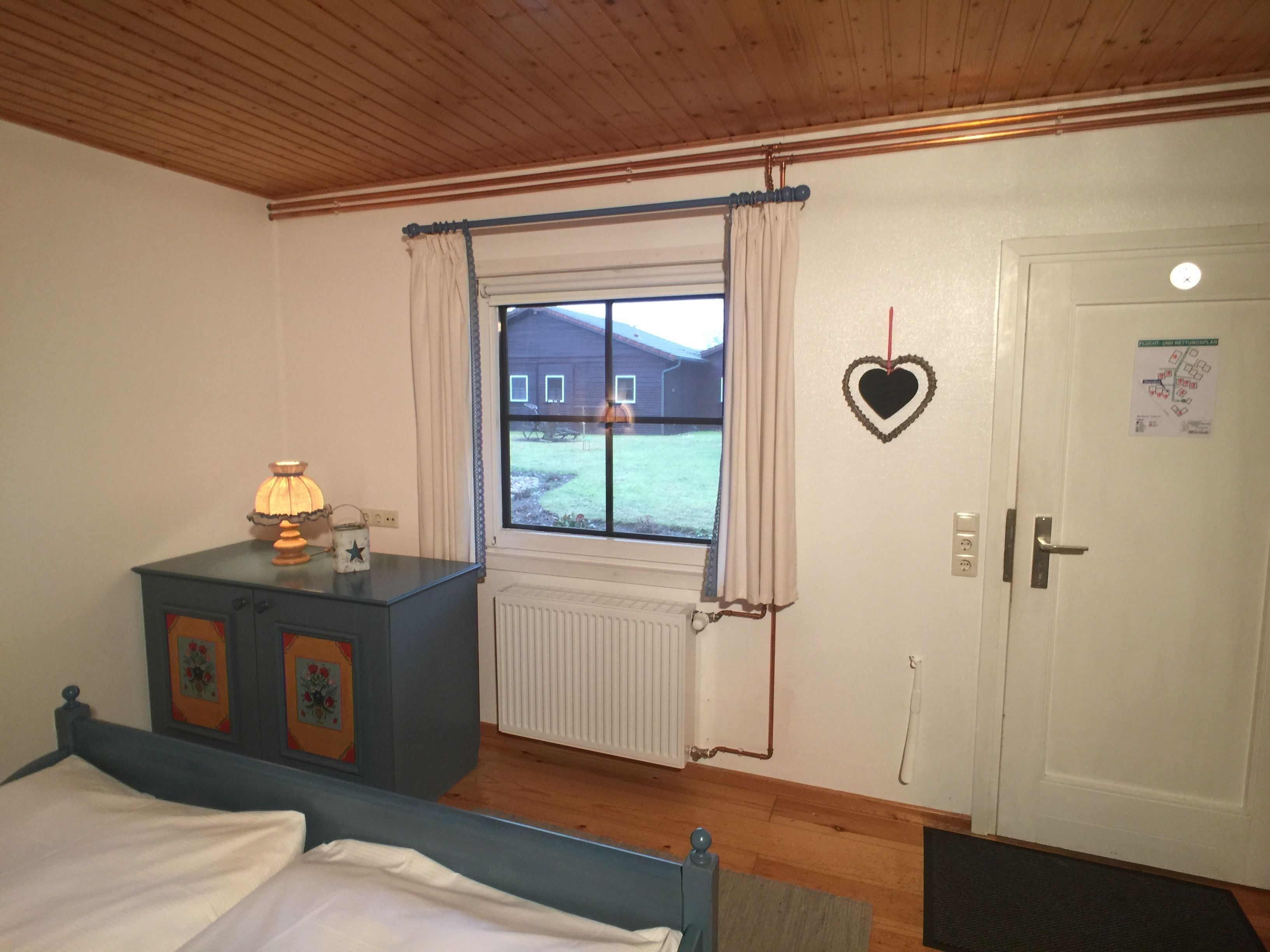 Un dormitorio con cama, cómoda, ventana con cortinas, radiador y puerta decorada con un corazón.