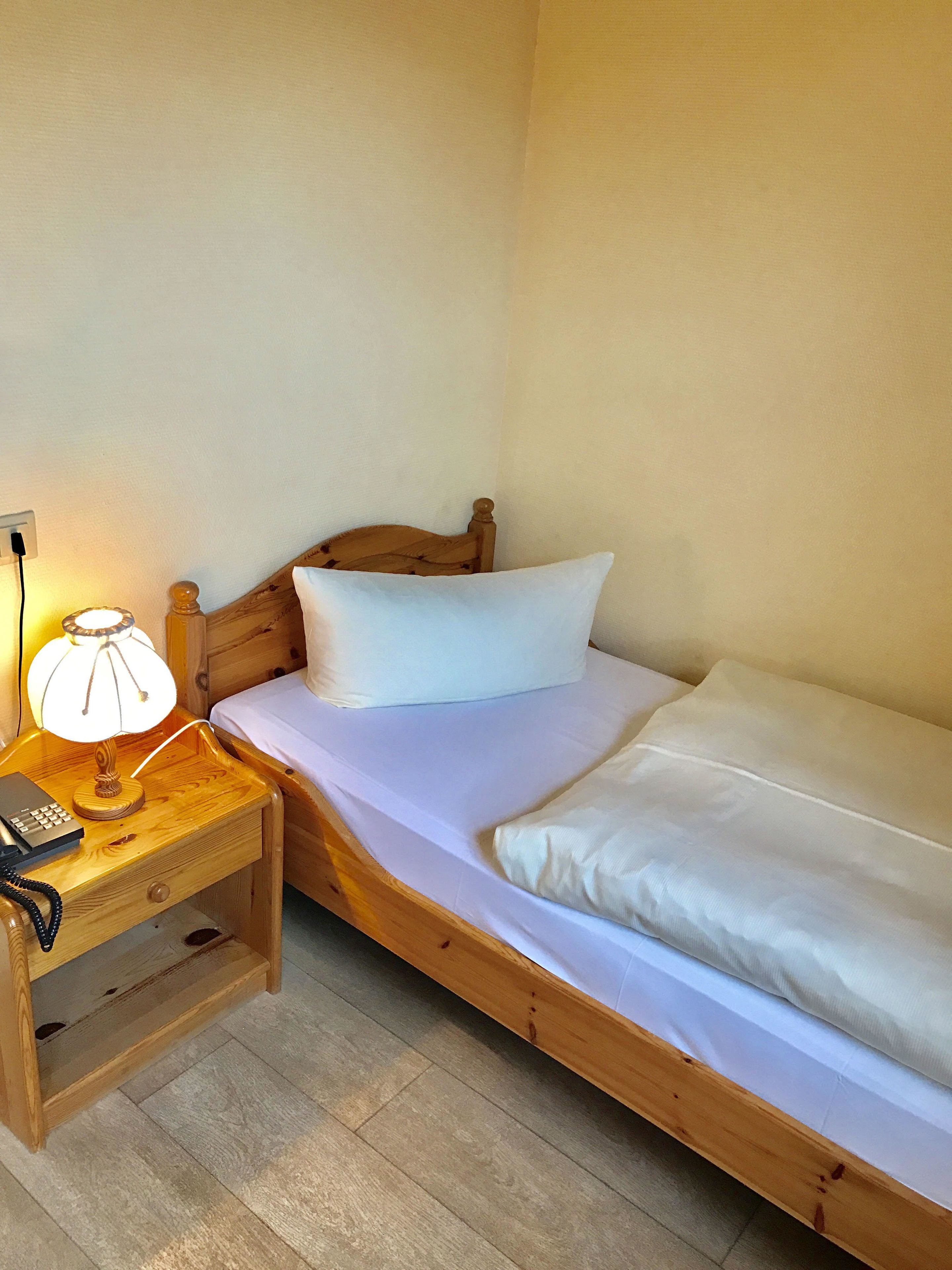 A single bed with a wooden headboard, a bedside table with a lamp, and a power outlet on the wall.