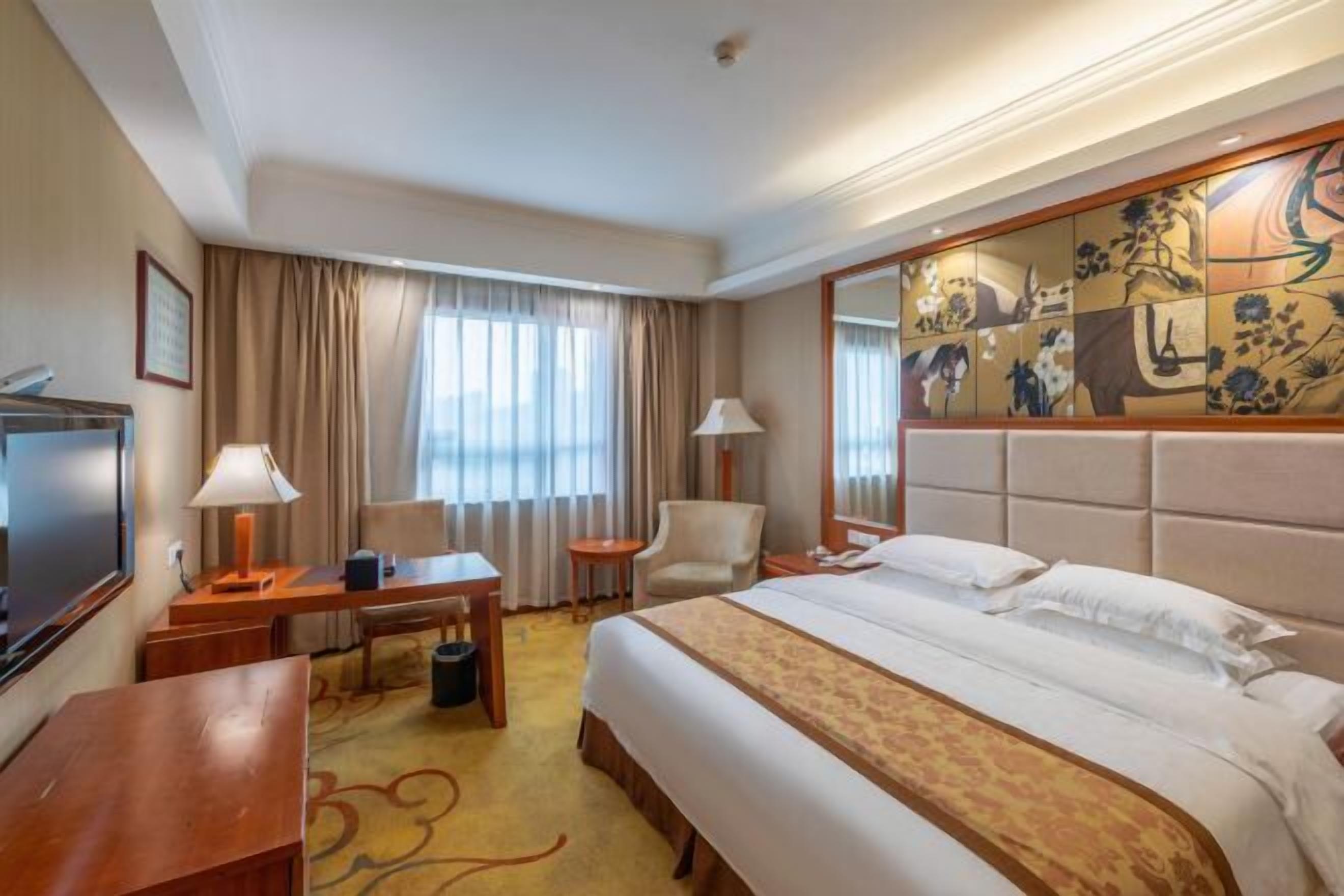 Habitación de hotel con una cama grande, un escritorio con lámpara, una silla, un televisor y un adorno en la pared.