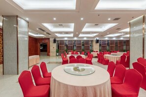 Un salón de banquetes con mesas redondas y sillas rojas, un techo alto con iluminación empotrada y vistas de la sala contigua a través de un panel de vidrio.