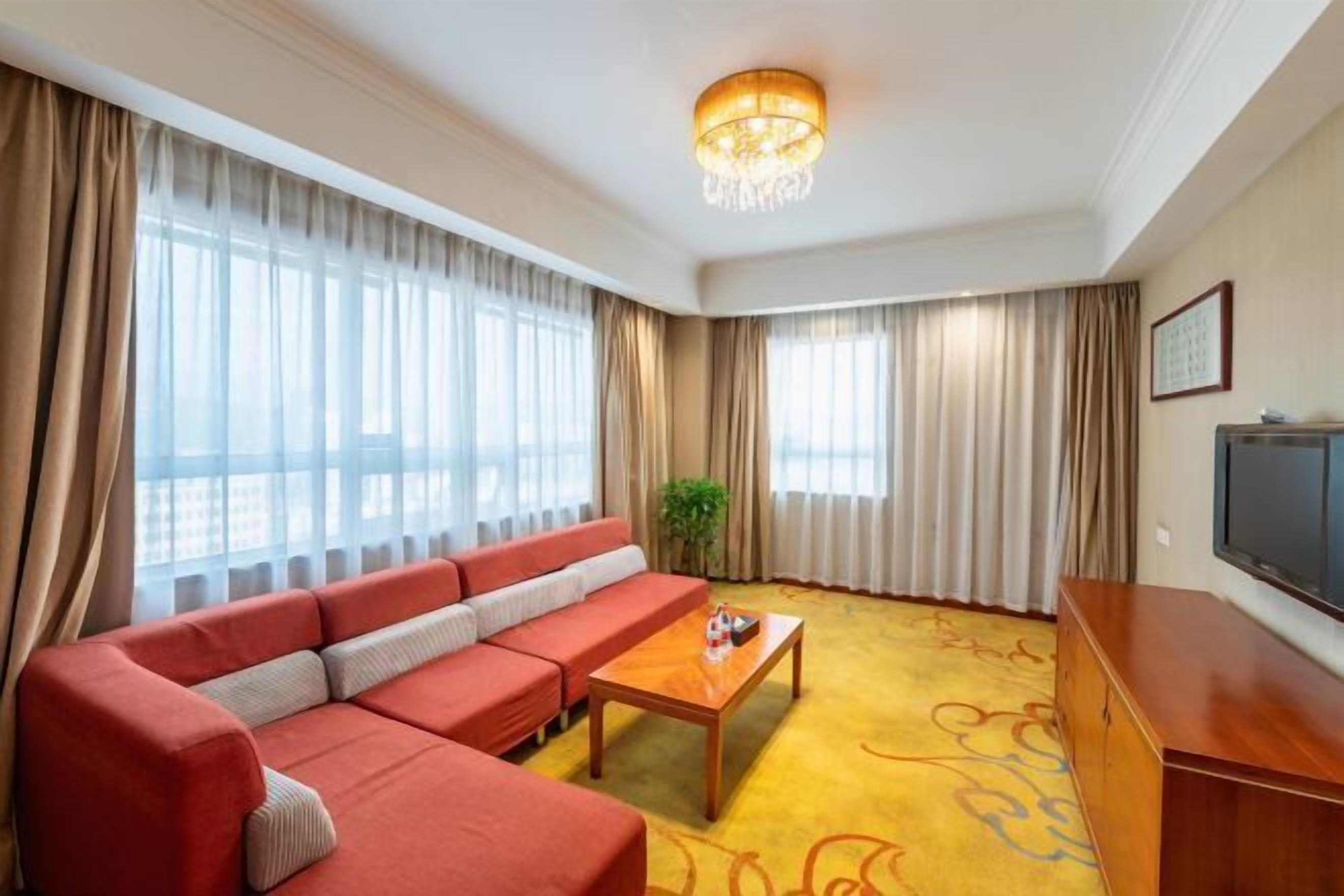 Ein Hotelzimmer mit einem roten Sofa, einem Holztisch, einem Fernseher und großen Fenstern mit Vorhängen.