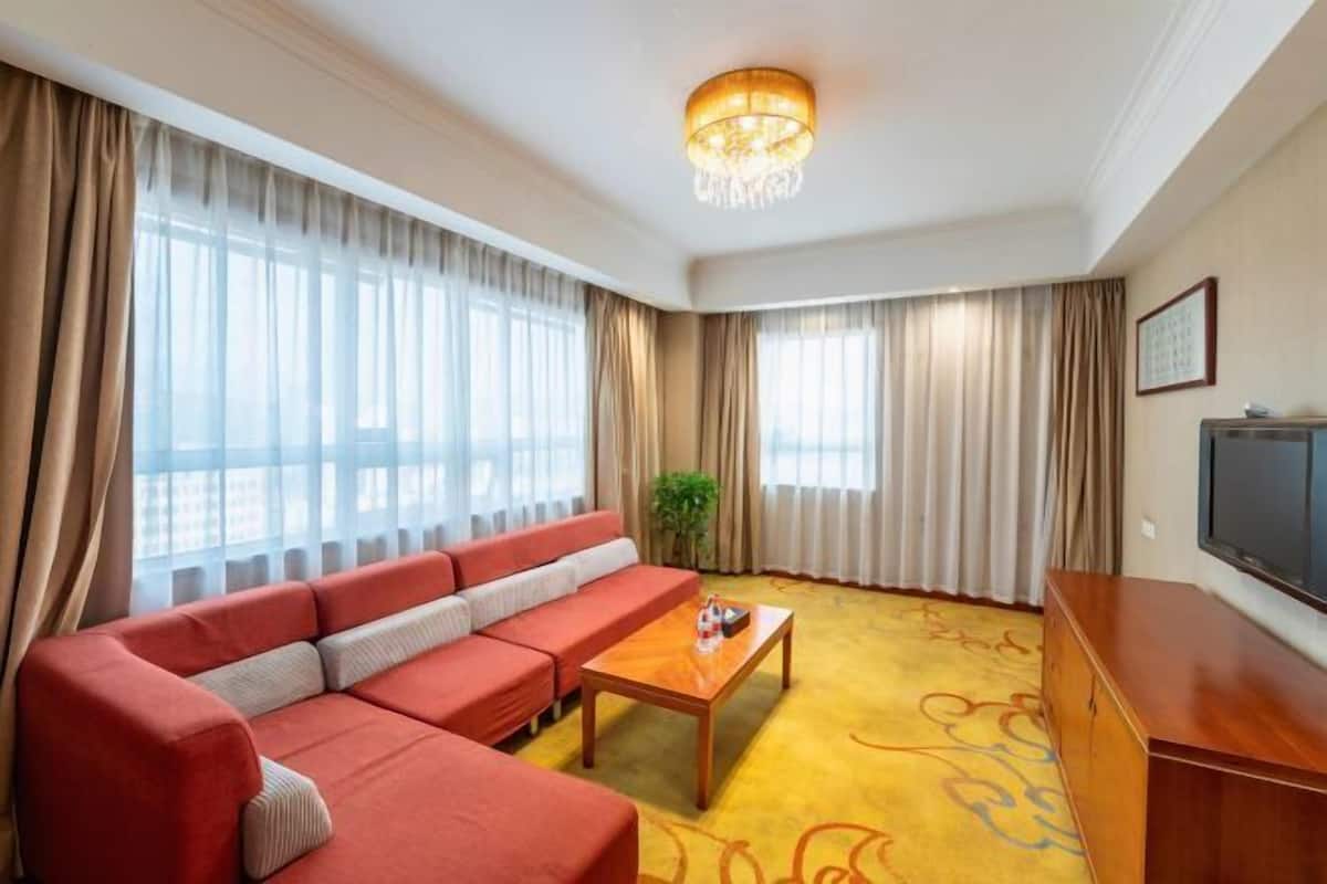 Una habitación de hotel con un sofá rojo, una mesa de centro de madera, un televisor y ventanas grandes con cortinas.