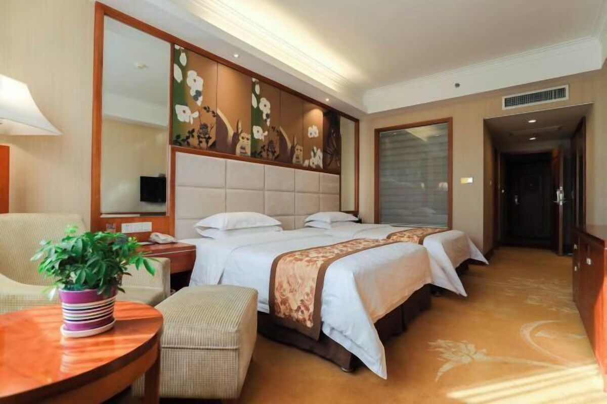A hotel room with a large bed, a desk, a chair, a potted plant, and a large mirror.