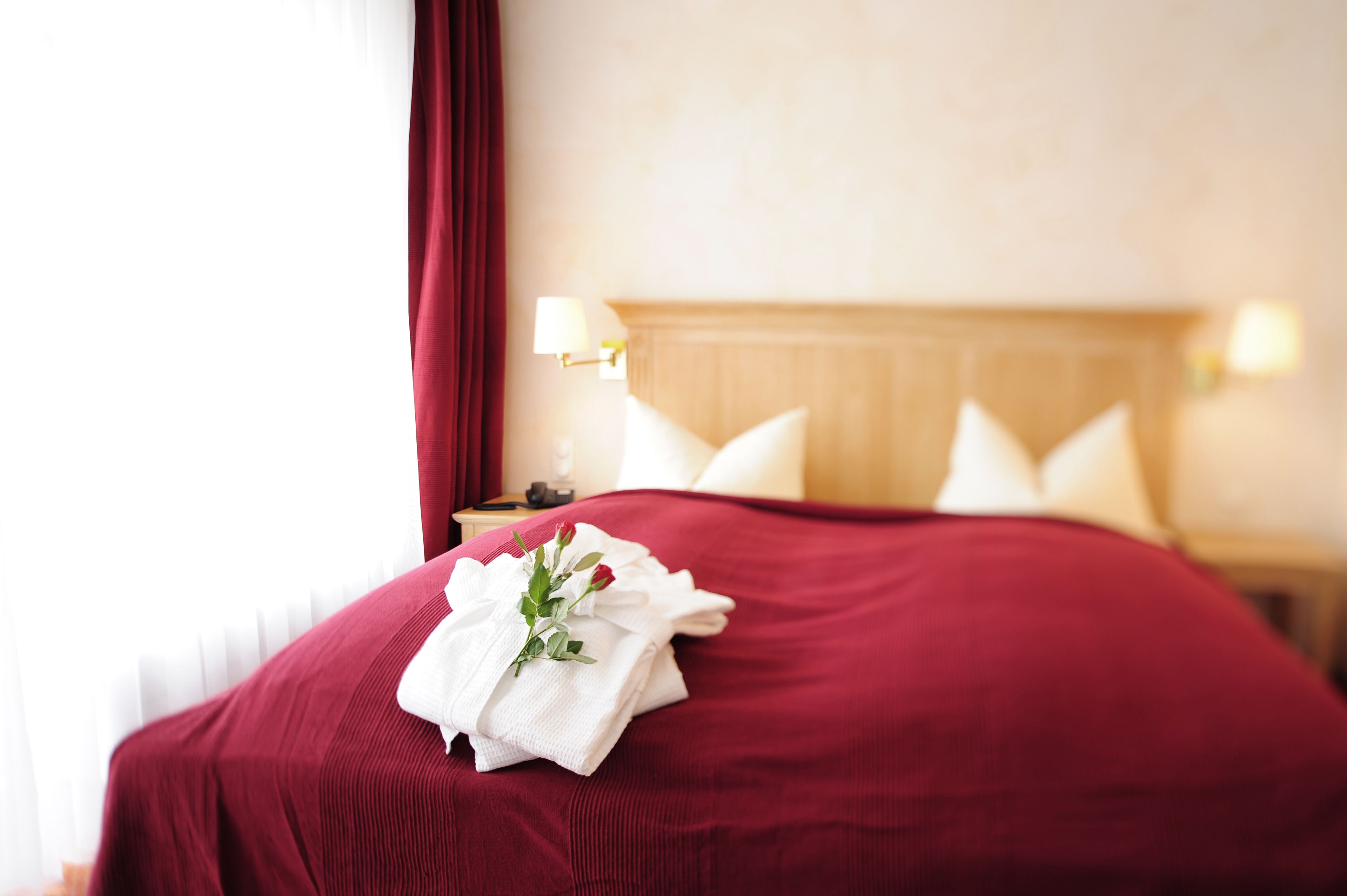 A neatly made bed with a red bedspread and white pillows, a white towel with a rose, and a nightstand with a lamp.