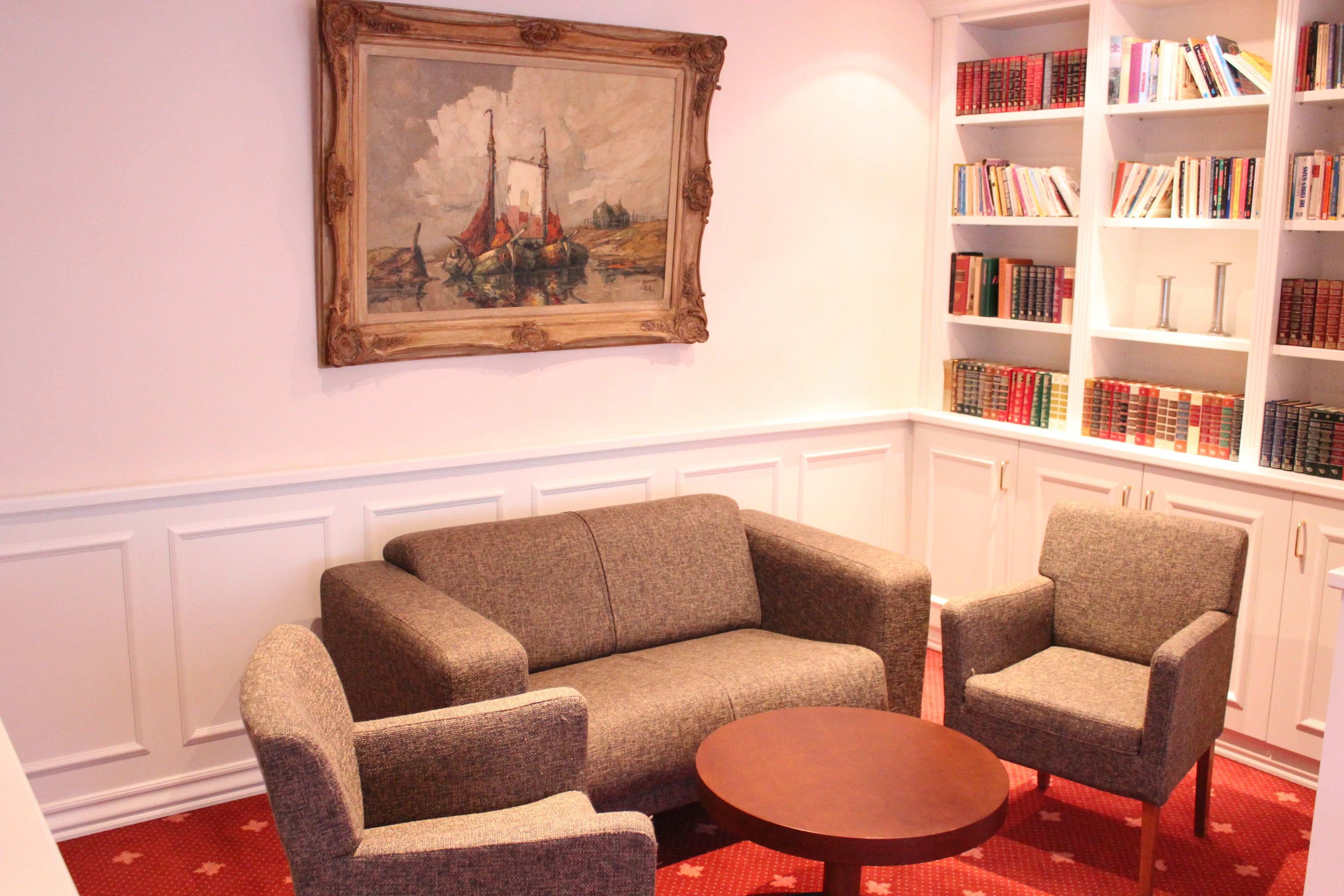 A room with a sofa, two armchairs, a small round table, a bookshelf, and a framed painting on the wall.