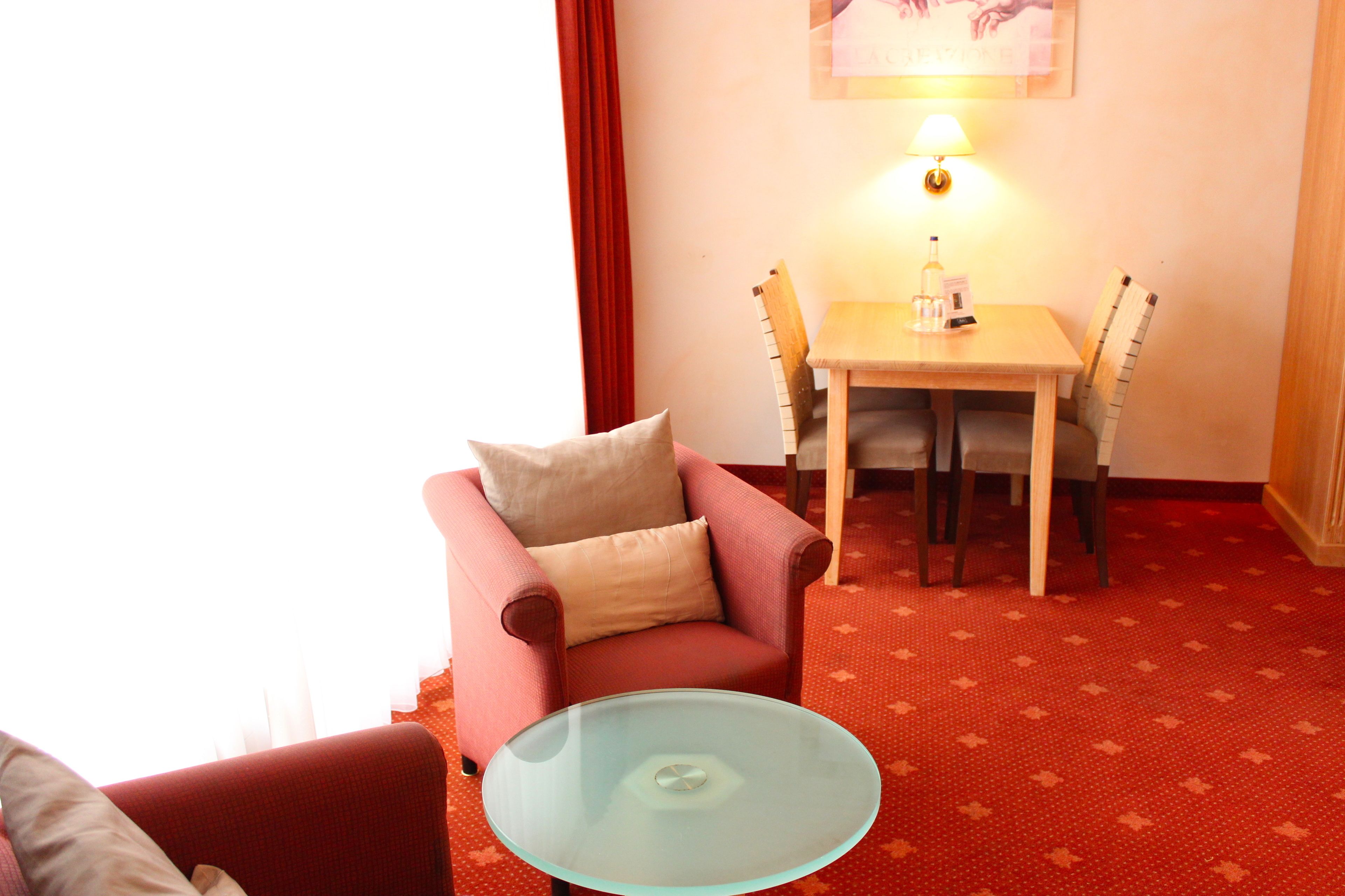 Una habitación con un sillón rojo, una mesa de vidrio, una mesa de comedor de madera y sillas, y una lámpara de pared.