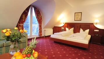 Ein Hotelzimmer mit Bett, Nachttisch, Tisch mit Obst und Blumen sowie einem Fenster mit Vorhängen.