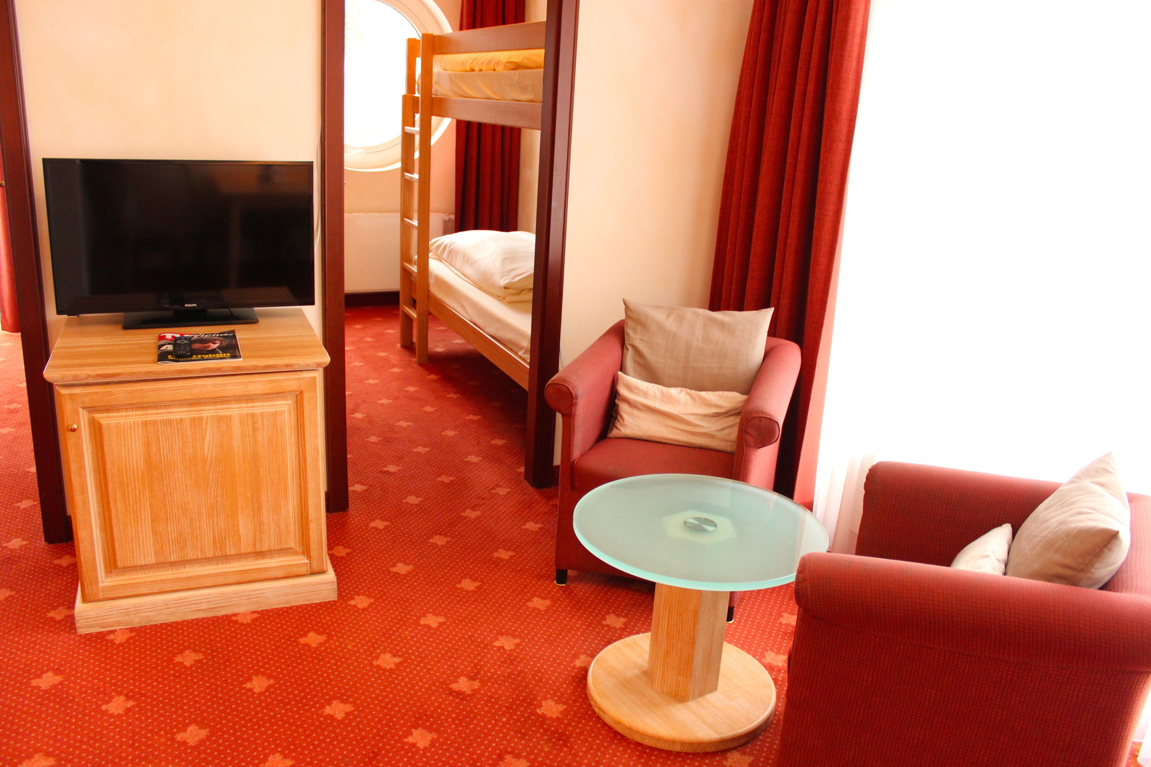 A hotel room with a television, bunk beds, a glass table, and red upholstered chairs.