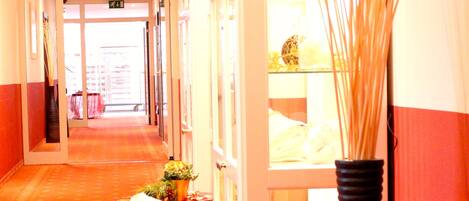 A hotel corridor with a reception desk, a black vase with reeds, and a patterned carpet.