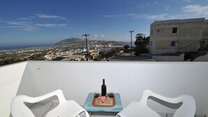 A terrace with two white chairs, a small table with a bottle and glasses, and a view of a town with buildings and a mountain in the distance. - King Thiras (Santorini)