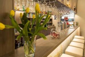 A bar area with a long counter, stools, a vase with yellow flowers, and shelves with bottles and glasses. - Sporthotel Wernigerode (Wernigerode)