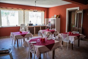 Ein Essbereich mit mehreren Tischen, gedeckt zum Essen, einer Küche im Hintergrund und großen Fenstern mit Vorhängen.