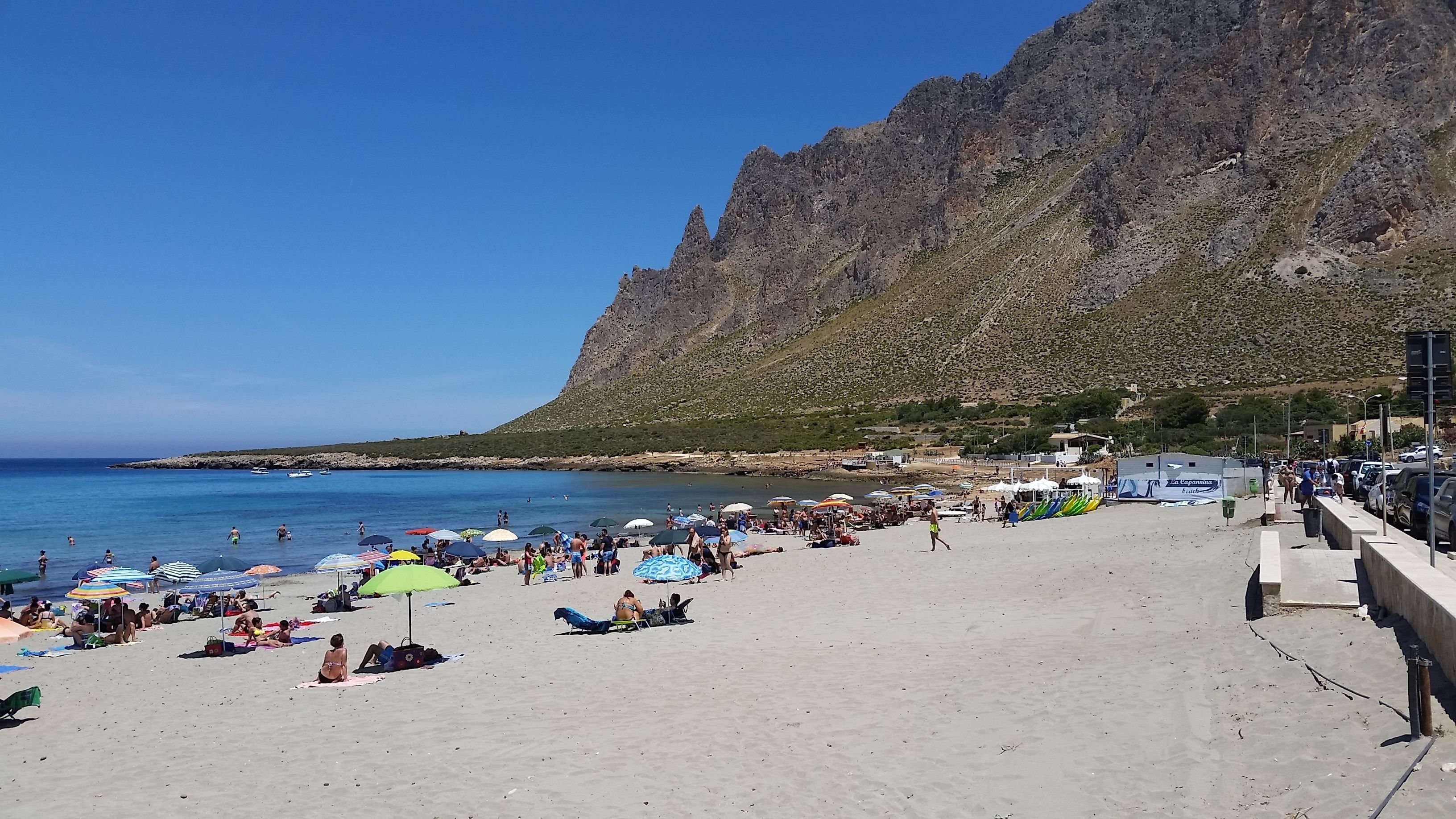 Ein überfüllter Strand mit badenden und sonnenden Personen, ein felsiger Klippen im Hintergrund und ein klarer, blauer Himmel.