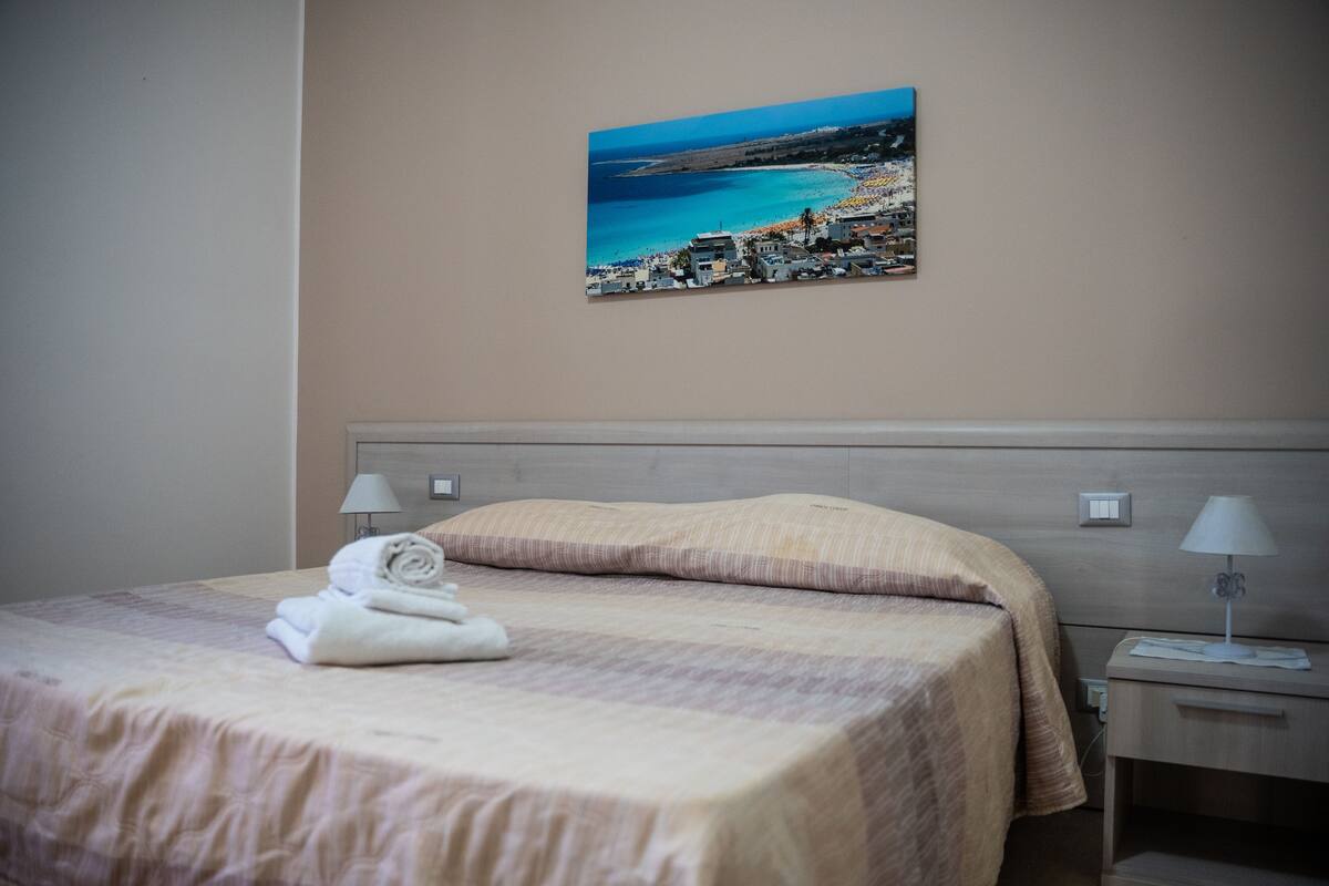 A neatly made bed with a folded towel, a headboard, bedside lamps, and a painting of a coastal scene on the wall.