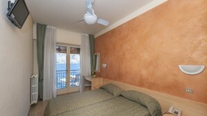 A hotel room with a bed, a desk, a chair, a television, a ceiling fan, and a view of the sea through a balcony door. - Hotel Sorriso (Brenzone sul Garda)