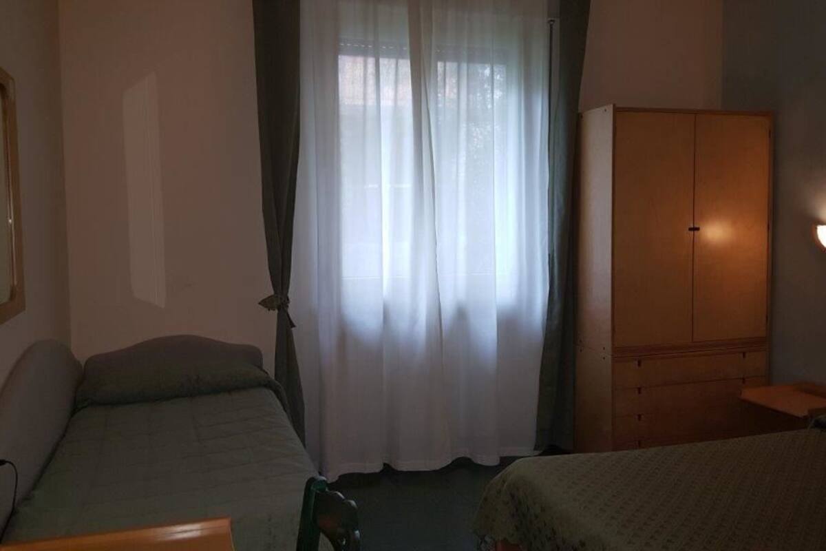 A hotel room with two single beds, a wooden wardrobe, a bedside table, and a wall-mounted light fixture.