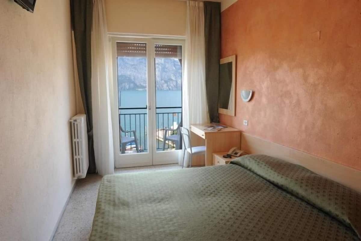 A hotel room with a bed, a desk, a chair, a phone, and a view of the sea through a sliding door.