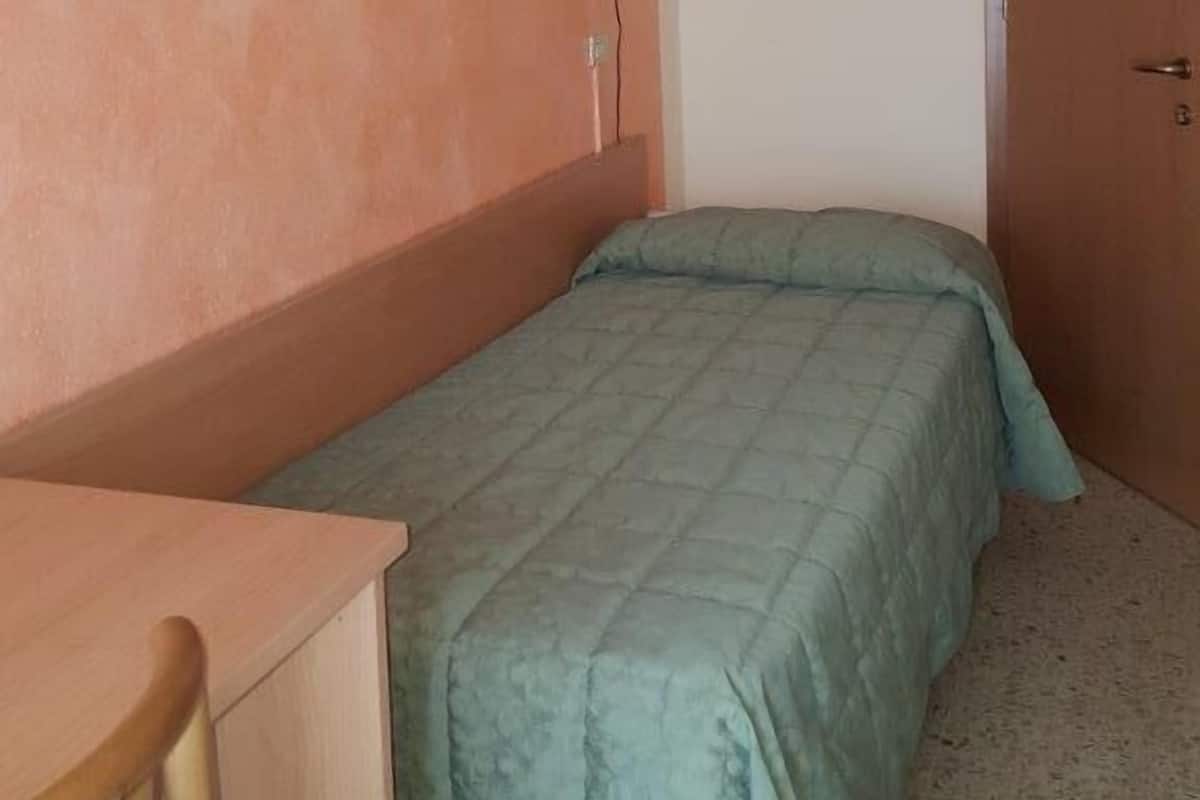 A small, single-bed room with a wooden desk and a chair, a wall-mounted lamp, and a door.
