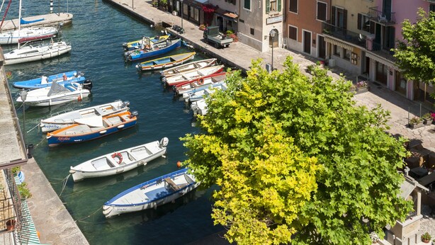 A marina with boats docked, buildings with balconies, and mountains in the background. - Hotel Sorriso (Brenzone sul Garda)