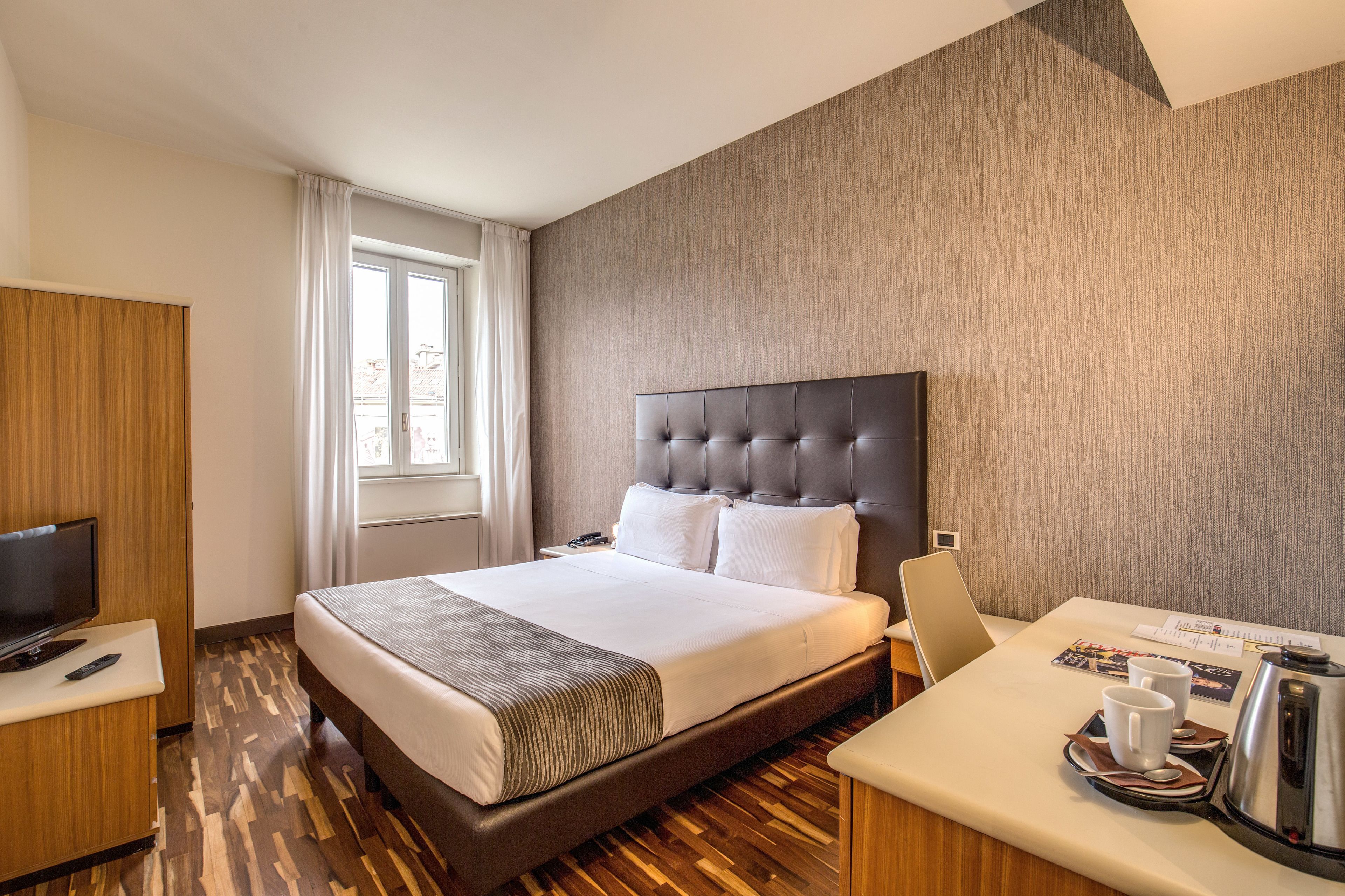 Habitación de hotel con una cama grande, televisión, escritorio con cafetera y ventana con cortinas.