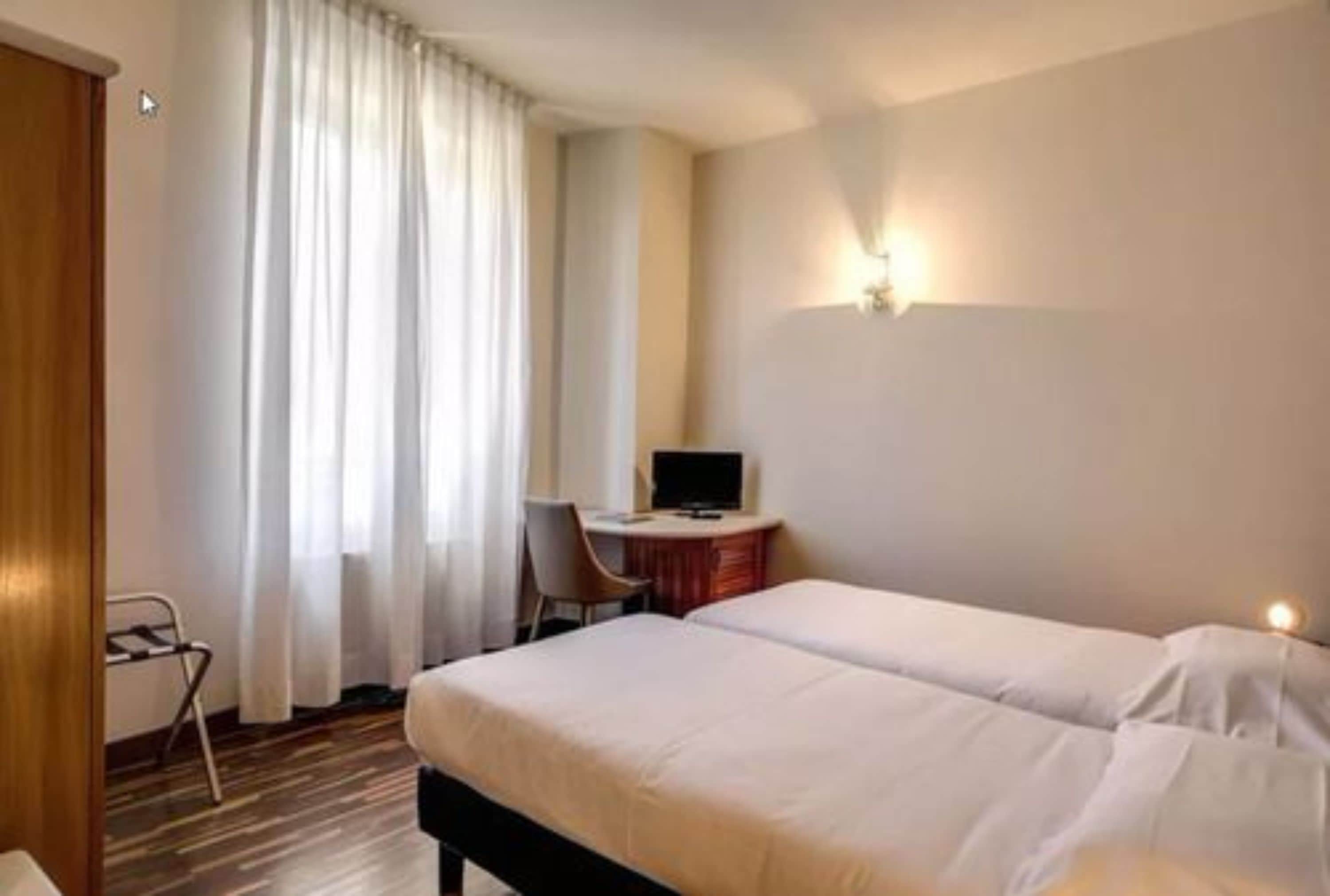 A hotel room with two single beds, a desk with a computer, a chair, and a window with curtains.