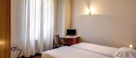 A hotel room with two single beds, a desk with a computer, a chair, and a window with curtains.