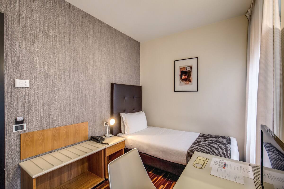 Habitación de hotel con cama, escritorio, silla, mesita de noche, lámpara, un cuadro en la pared y ventana con cortinas.