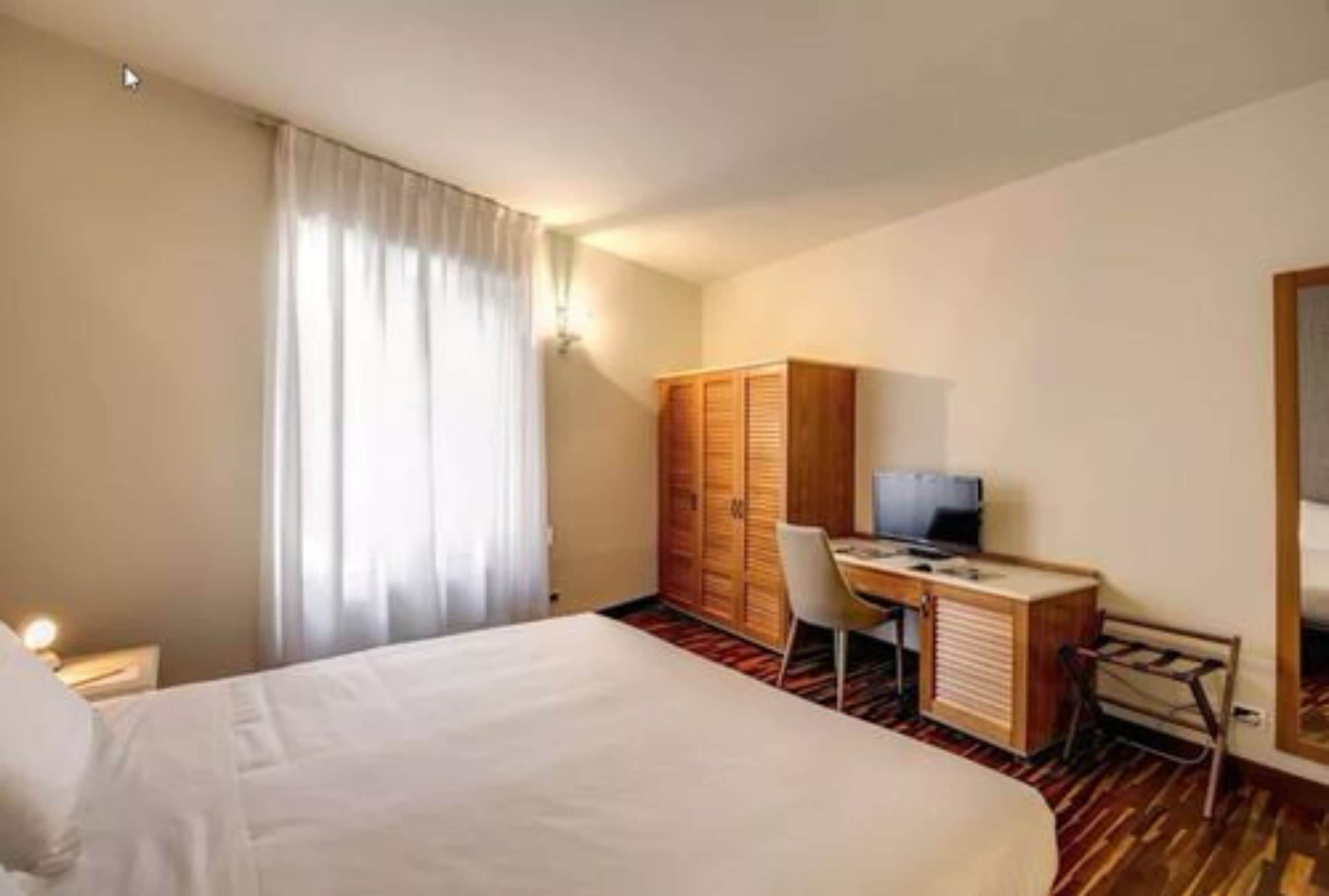 A hotel room with a bed, a desk, a chair, a wardrobe, and a television.