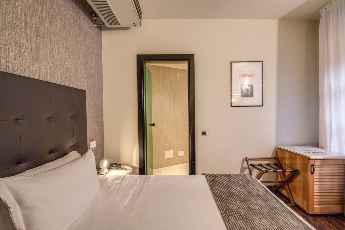 Habitación de hotel con una cama grande, una mesita de noche con lámpara, un escritorio de madera pequeño y un cuadro en la pared.