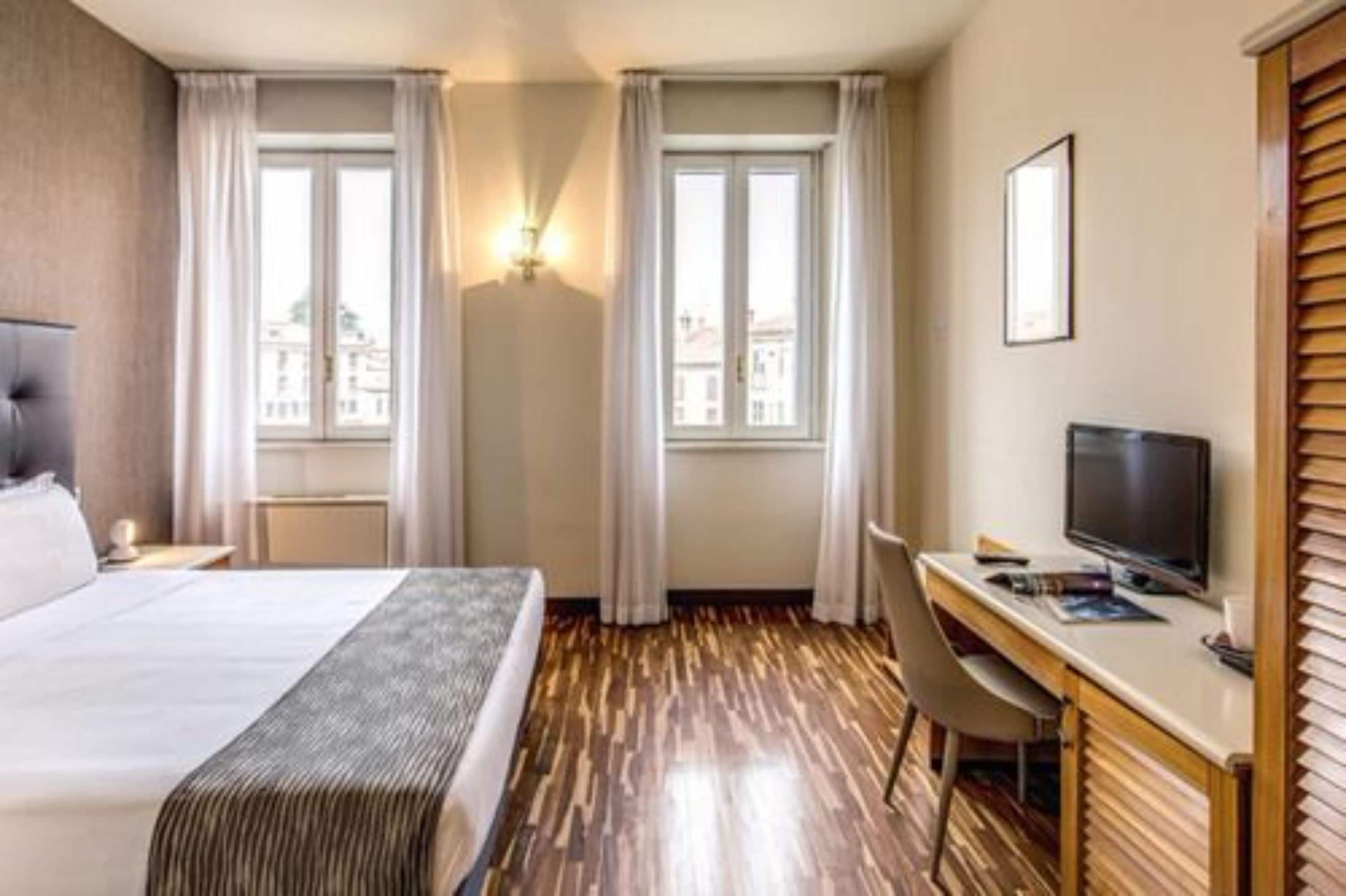 A hotel room with a bed, a desk with a computer, two windows with curtains, and a television.