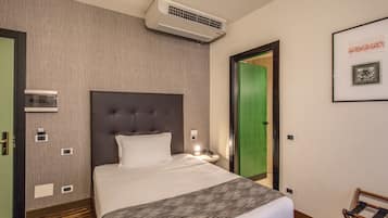 A hotel room with a large bed, a wall-mounted air conditioner, a bedside table with a lamp, and a framed picture on the wall.