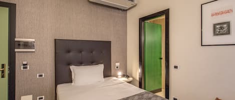 A hotel room with a large bed, a wall-mounted air conditioner, a bedside table with a lamp, and a framed picture on the wall.