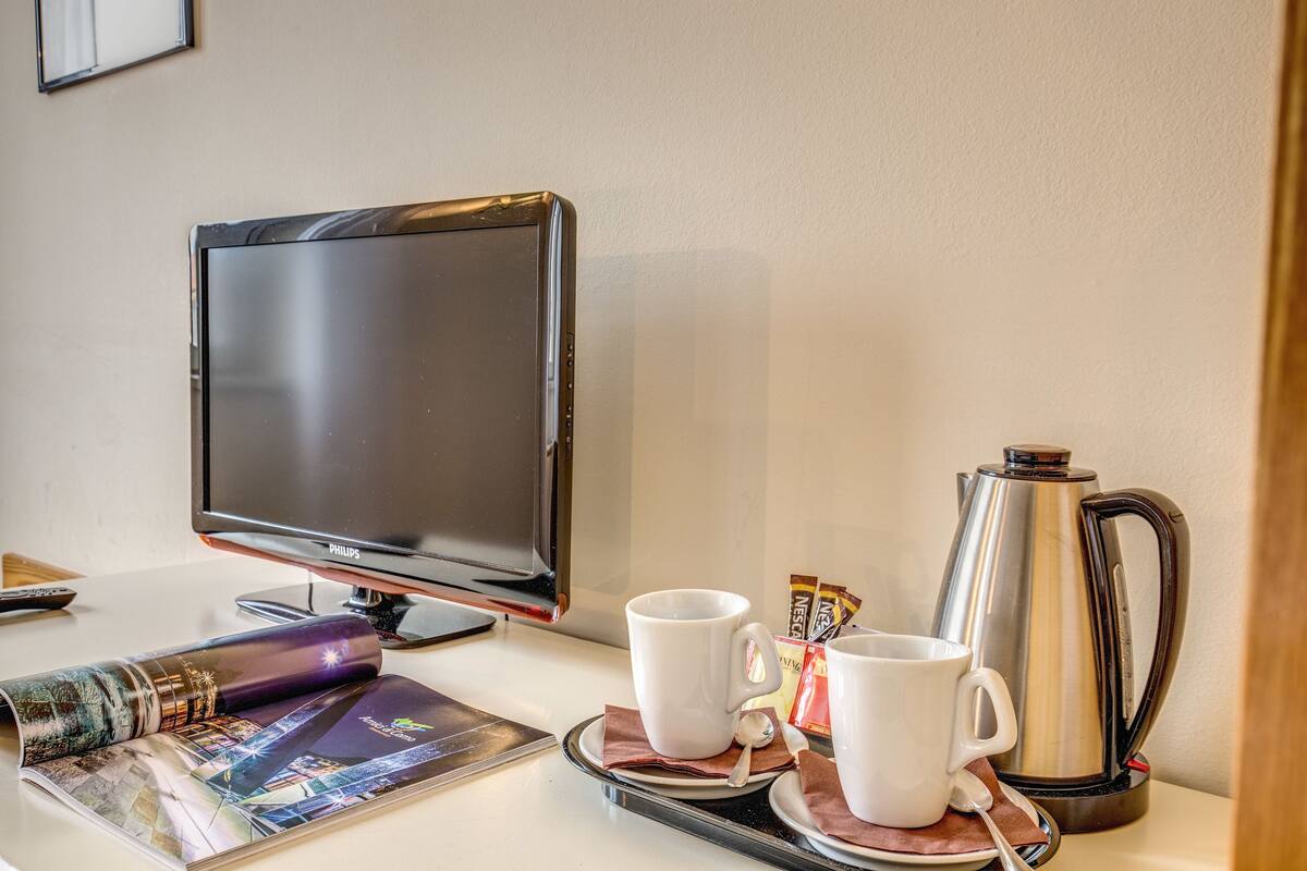 Habitación de hotel con televisión de pantalla plana, dos tazas blancas, cafetera y una revista sobre la mesa.