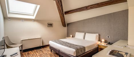 A bedroom with a bed, a desk, a chair, a bedside table, a lamp, and a skylight.