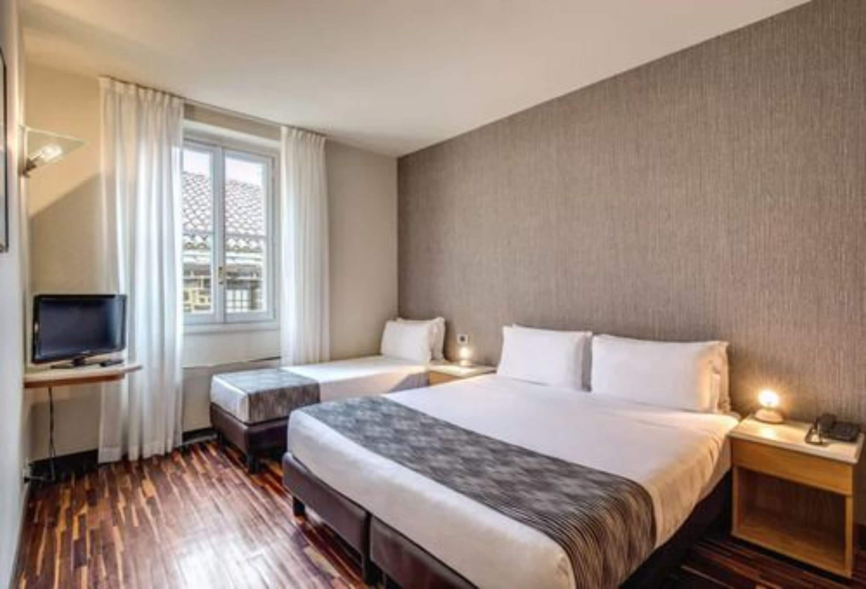 Habitación de hotel con dos camas, televisión, ventana con cortinas y piso de madera.