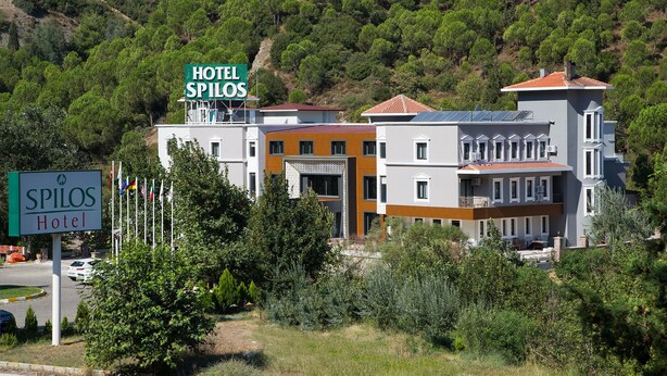 Um hotel chamado SPILOS Hotel, com uma placa, rodeado por vegetação e edifícios modernos.