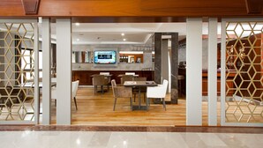 Um lobby moderno de hotel com um balcão de recepção de madeira, uma área de jantar com mesas e cadeiras, e um bar visível através de um painel decorado.