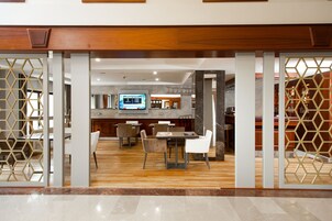 Un moderno vestíbulo de hotel con un mostrador de recepción de madera, un área de comedor con mesas y sillas, y una zona de bar visible a través de un separador decorativo.