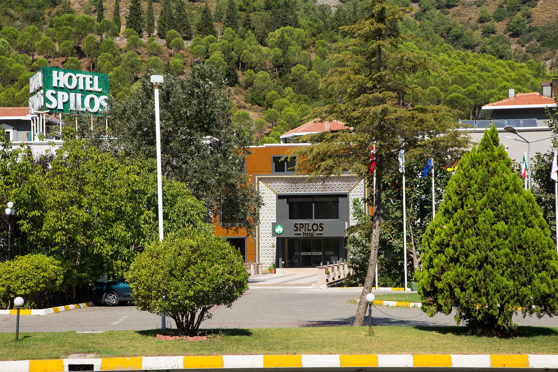 Um hotel chamado 'Hotel Spilos', com fachada moderna, rodeado por vegetação e uma paisagem montanhosa.