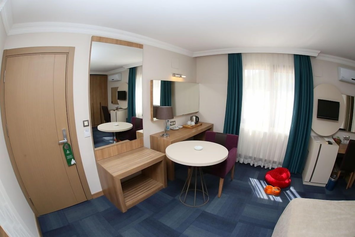 A hotel room with a bed, a desk, a chair, a small table, a mirror, a TV, and a window with curtains.
