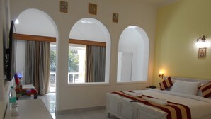 A hotel room with a bed, a desk, and a chair. There are three windows and a door. - Hotel Isabel Palace (Rajnagar)