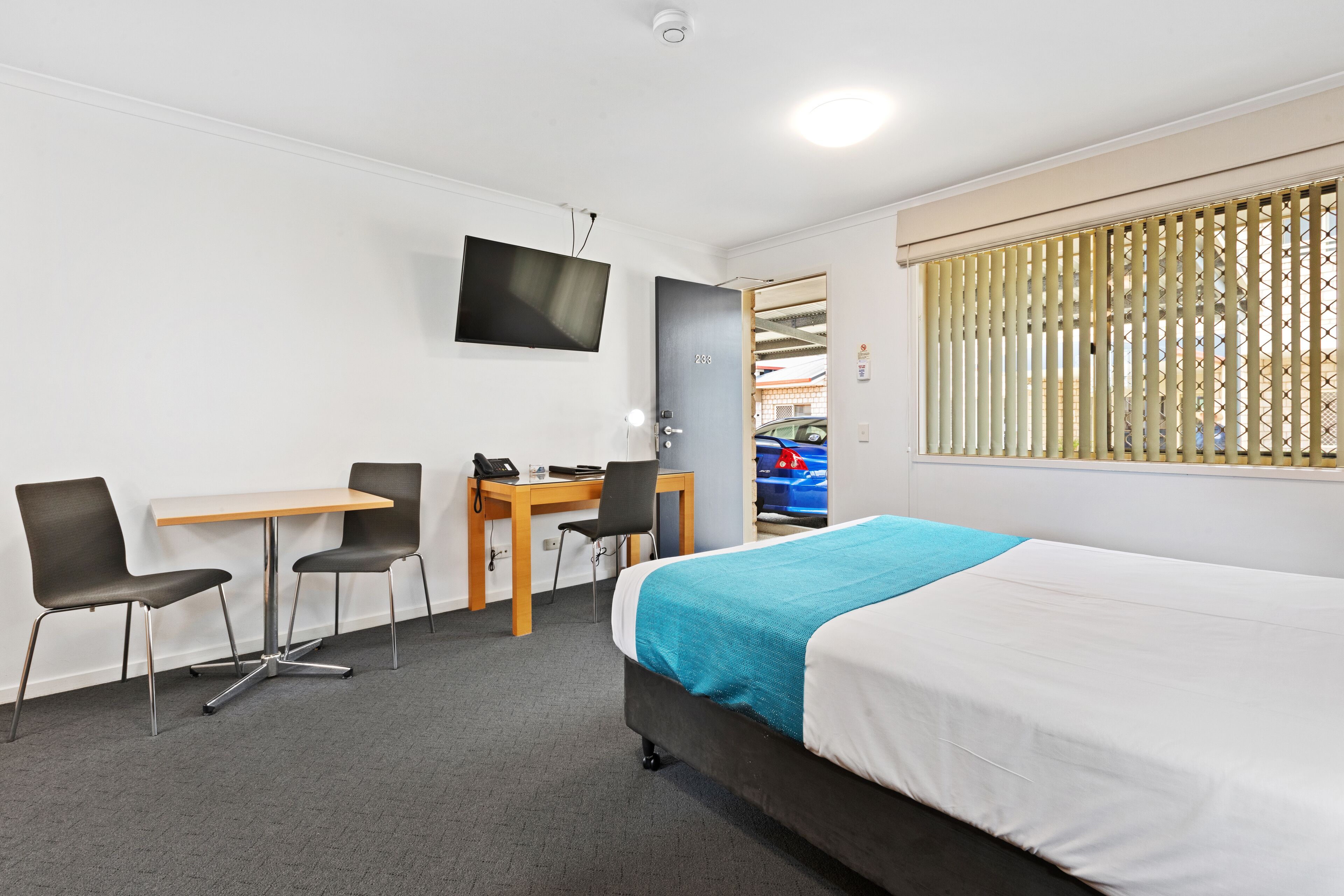 A hotel room with a bed, a desk with a chair, a TV mounted on the wall, and a window with blinds.