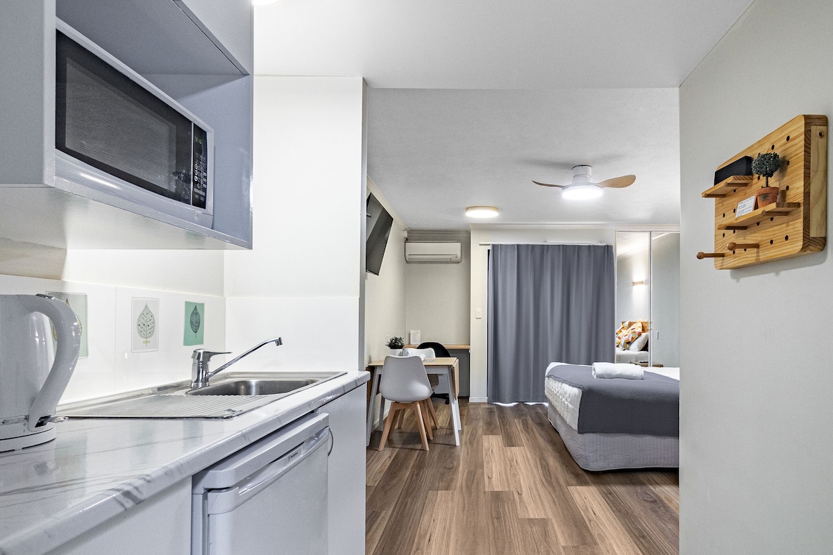 A compact kitchen with a microwave, sink, and modern appliances. A dining area with a table and chairs is visible, leading to a bedroom with a bed and a ceiling fan.