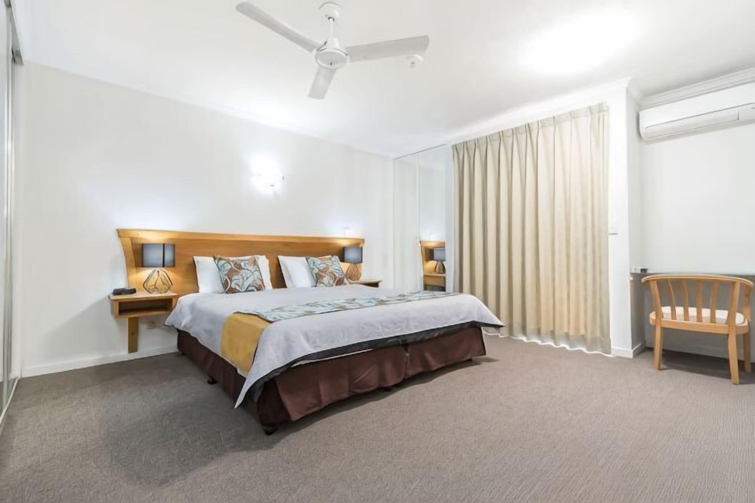 A hotel room with a bed, bedside lamps, a ceiling fan, a chair, and a large window with curtains.
