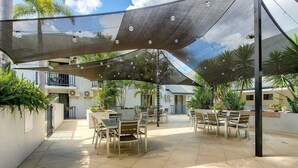 An outdoor patio area with tables and chairs, shaded by a large canopy, surrounded by greenery and buildings. - Parkview Apartments (South Brisbane)