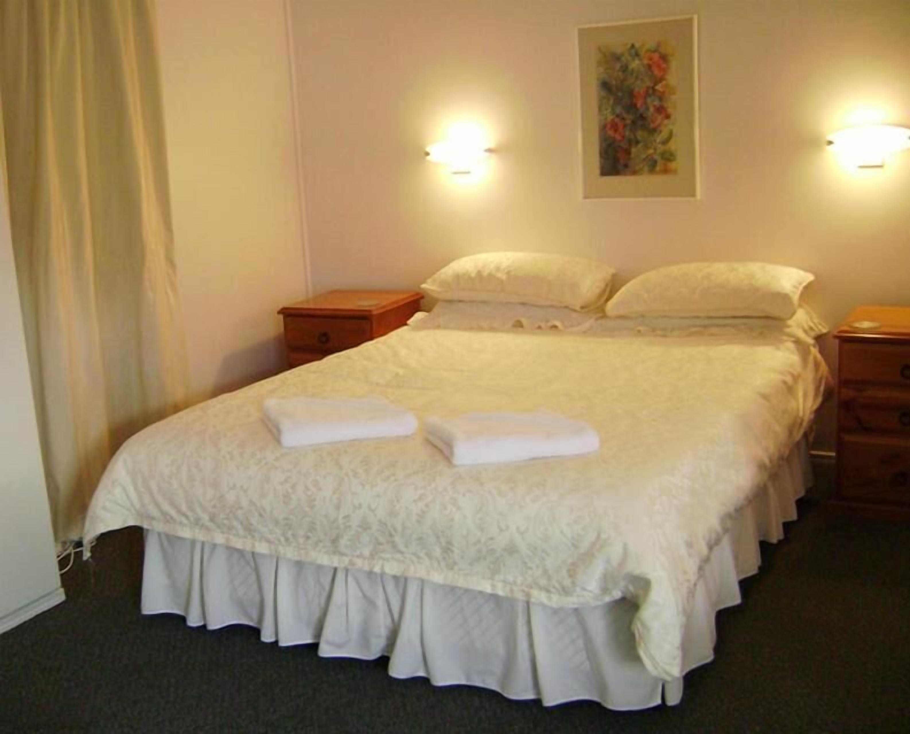 Una cama bien hecha con una colcha blanca y dos toallas dobladas, una mesita de noche con una lámpara y un cuadro en la pared.