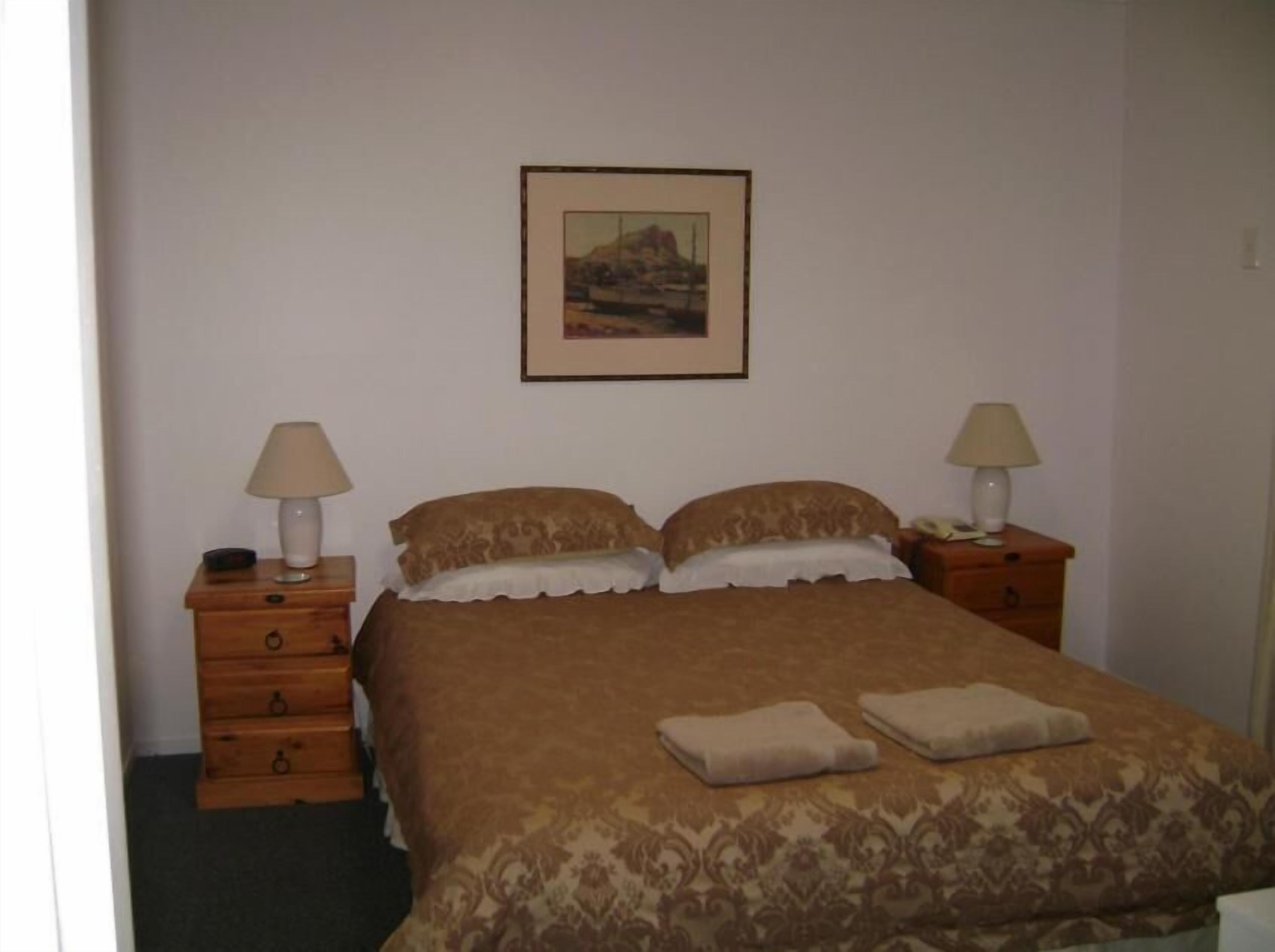 A bedroom with a bed, two nightstands, a lamp on each, a framed picture on the wall, and a carpeted floor.