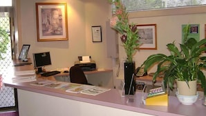 A reception desk with a computer, a telephone, and a plant. There are framed pictures on the wall and a window with blinds. - River Park Motor Inn (Casino)
